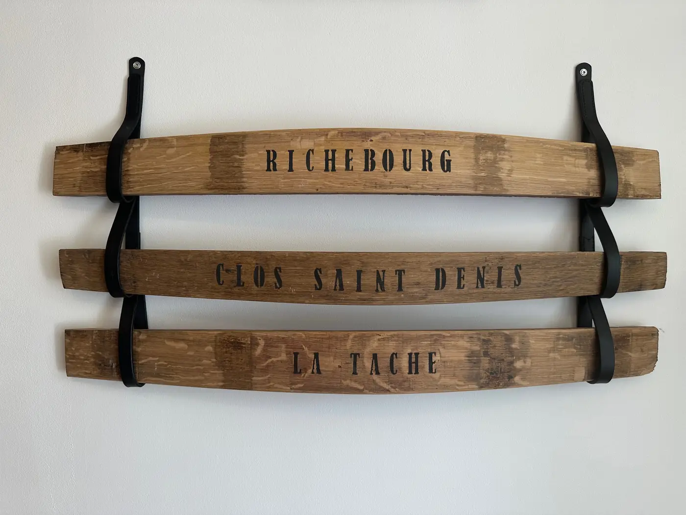 Trois douelles de tonneau peintes Richebourg, Clos Saint-Denis et La Tâche, suspendues au mur par une paire d’attaches en cuir à trois boucles