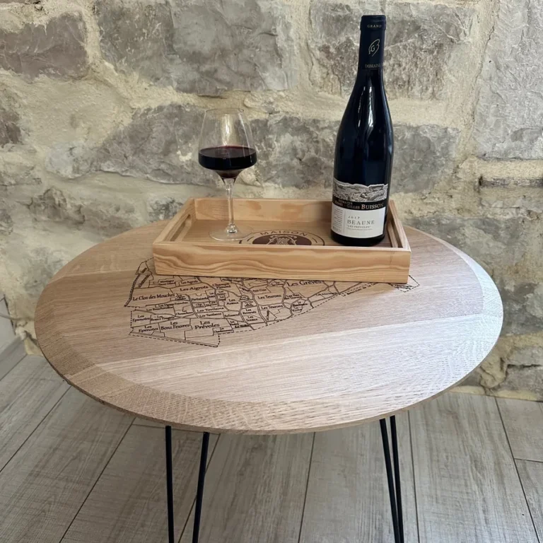 Table basse ronde en chêne gravée d’une carte viticole, pieds épingle noirs, devant un mur en pierre