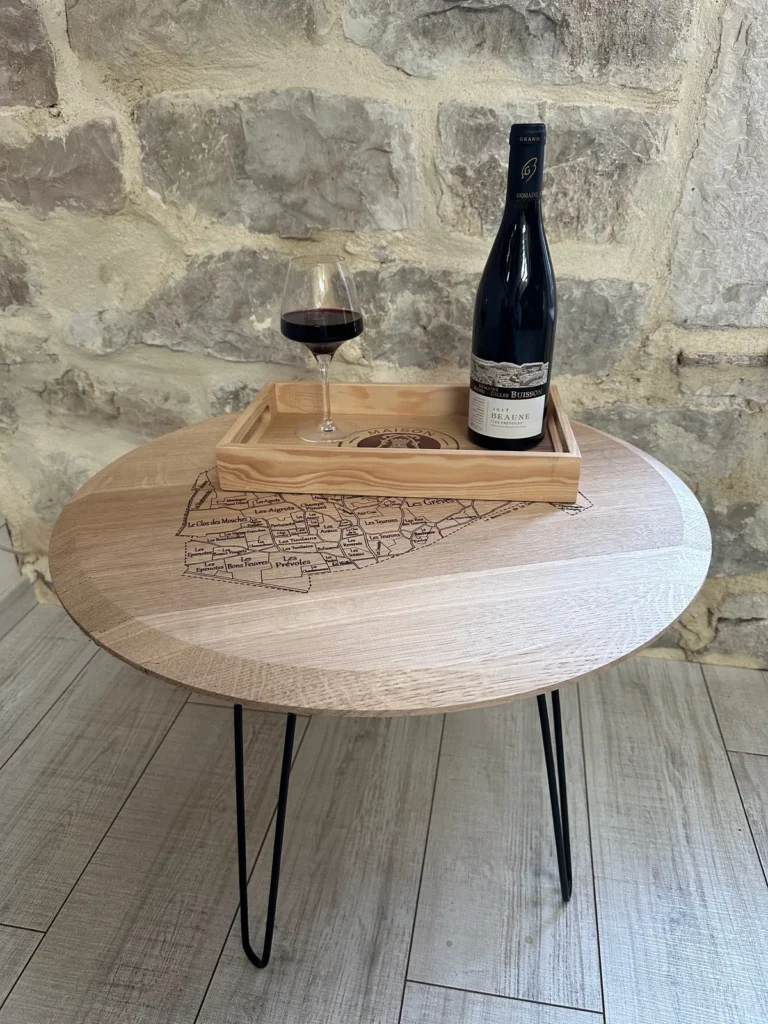 Table basse ronde en chêne gravée d’une carte viticole, pieds épingle noirs, devant un mur en pierre